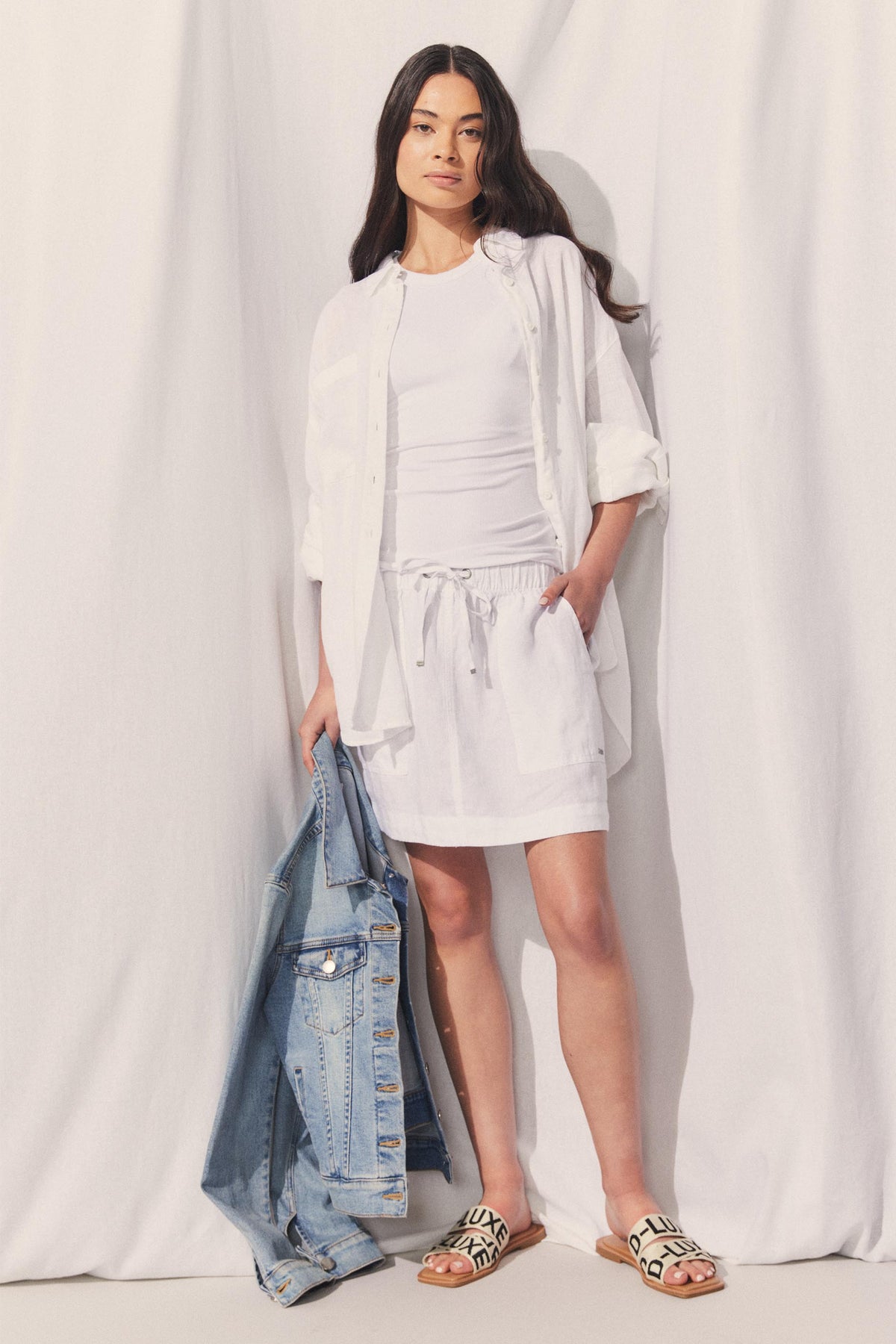 Model wearing D-Luxe Linen Mini Skirt with elastic waistband and side pockets, holding denim jacket in portrait view.