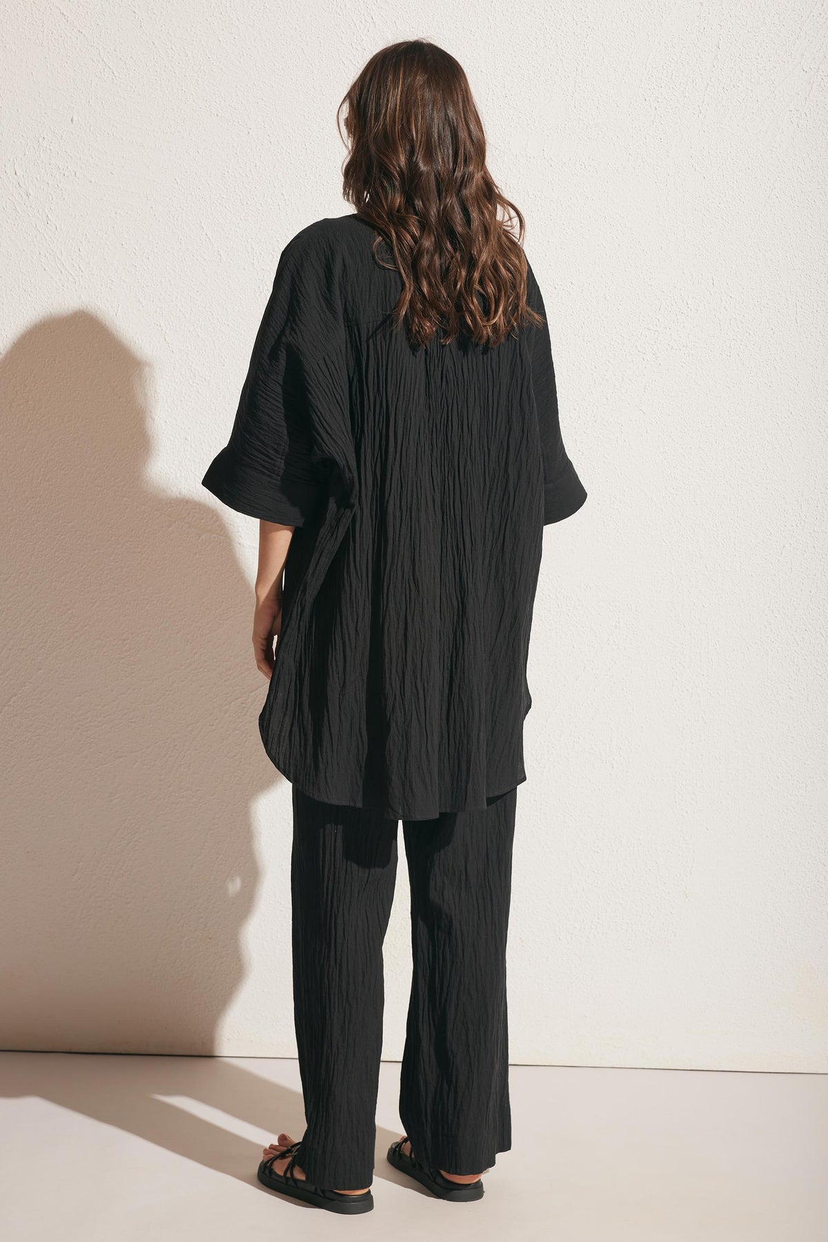 Woman seen from behind wearing a relaxed black textured long shirt and wide-leg pants with sandals against a light wall
