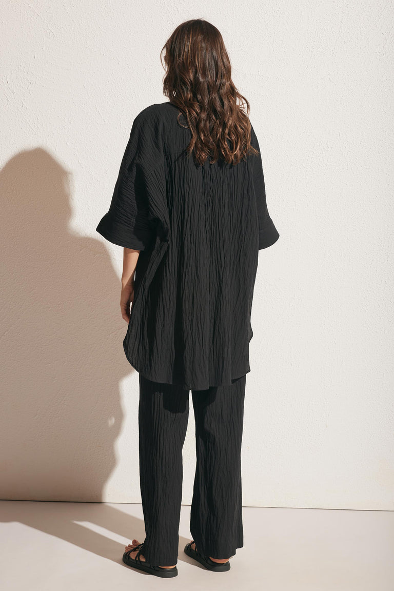 Woman seen from behind wearing a relaxed black textured long shirt and wide-leg pants with sandals against a light wall