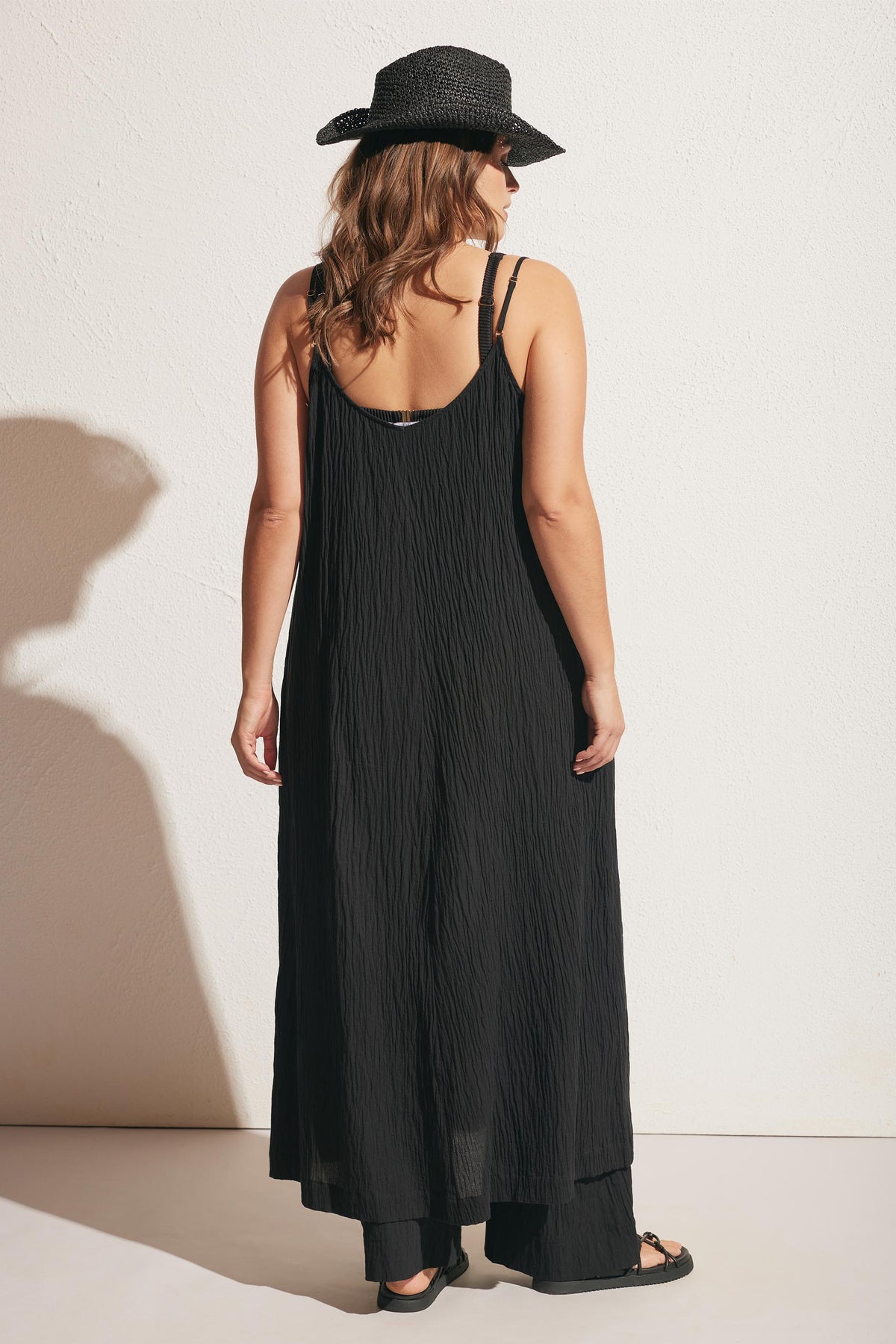 Woman wearing a textured black maxi dress with thin straps, paired with a black woven hat and sandals, standing against white wall