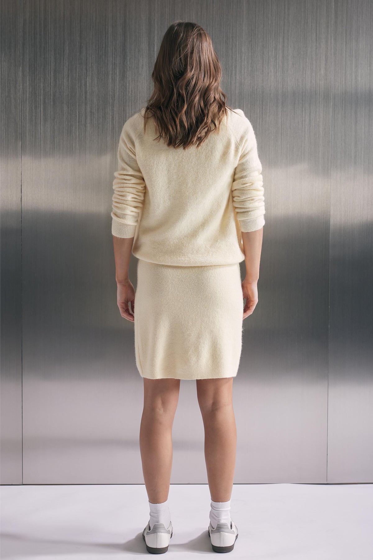 Woman wearing a soft cream knit mini skirt paired with a matching relaxed sweater and white sneakers, back view