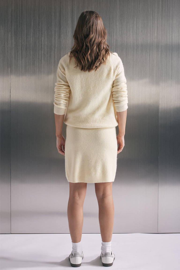 Woman wearing a soft cream knit mini skirt paired with a matching relaxed sweater and white sneakers, back view