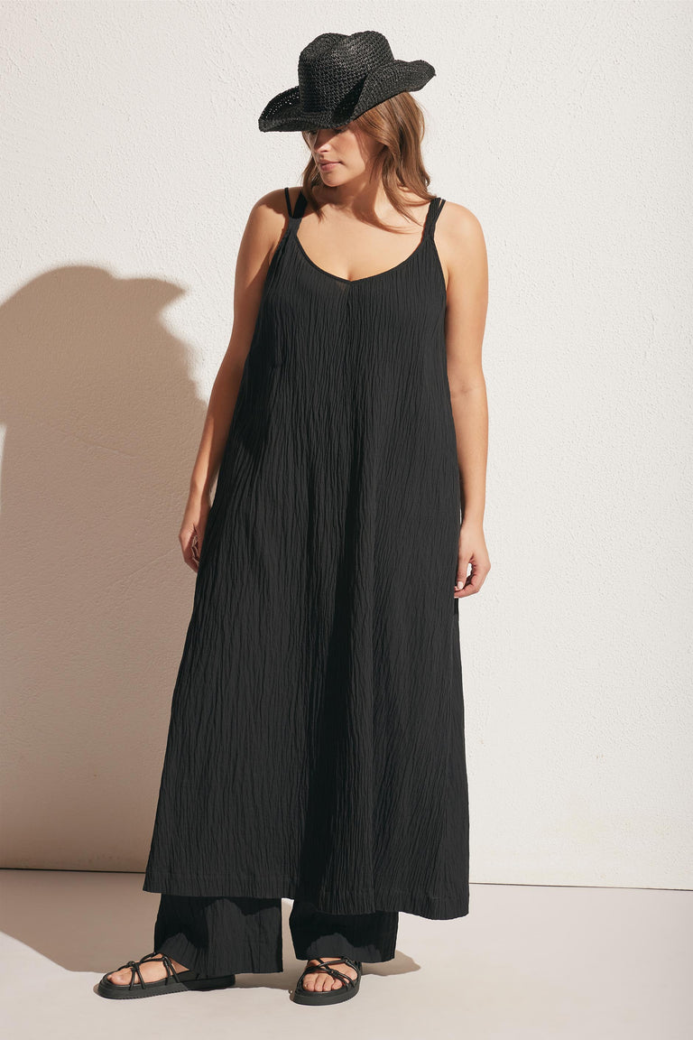Woman wearing a textured black sleeveless maxi dress layered over wide-leg pants with a woven black hat and sandals