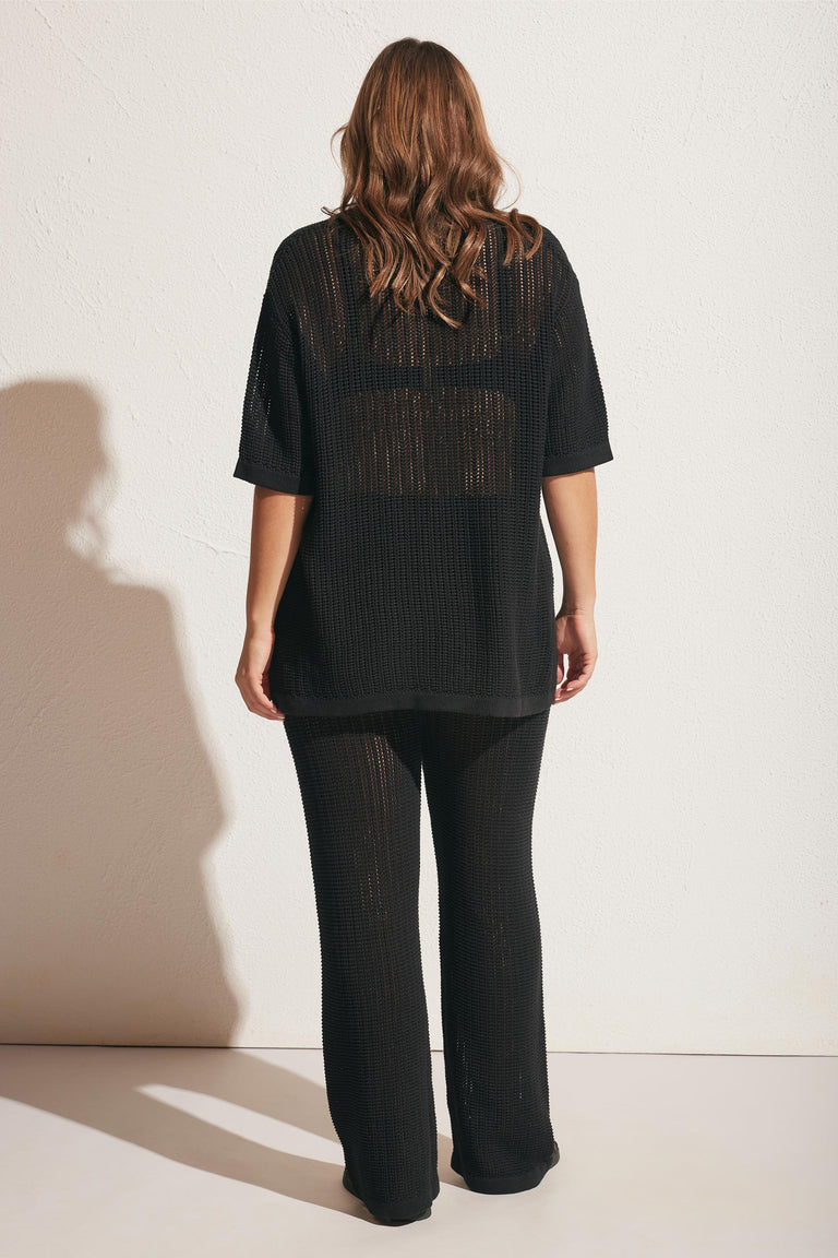 Woman wearing loose black crochet pants and matching oversized crochet top, standing against a light wall from behind