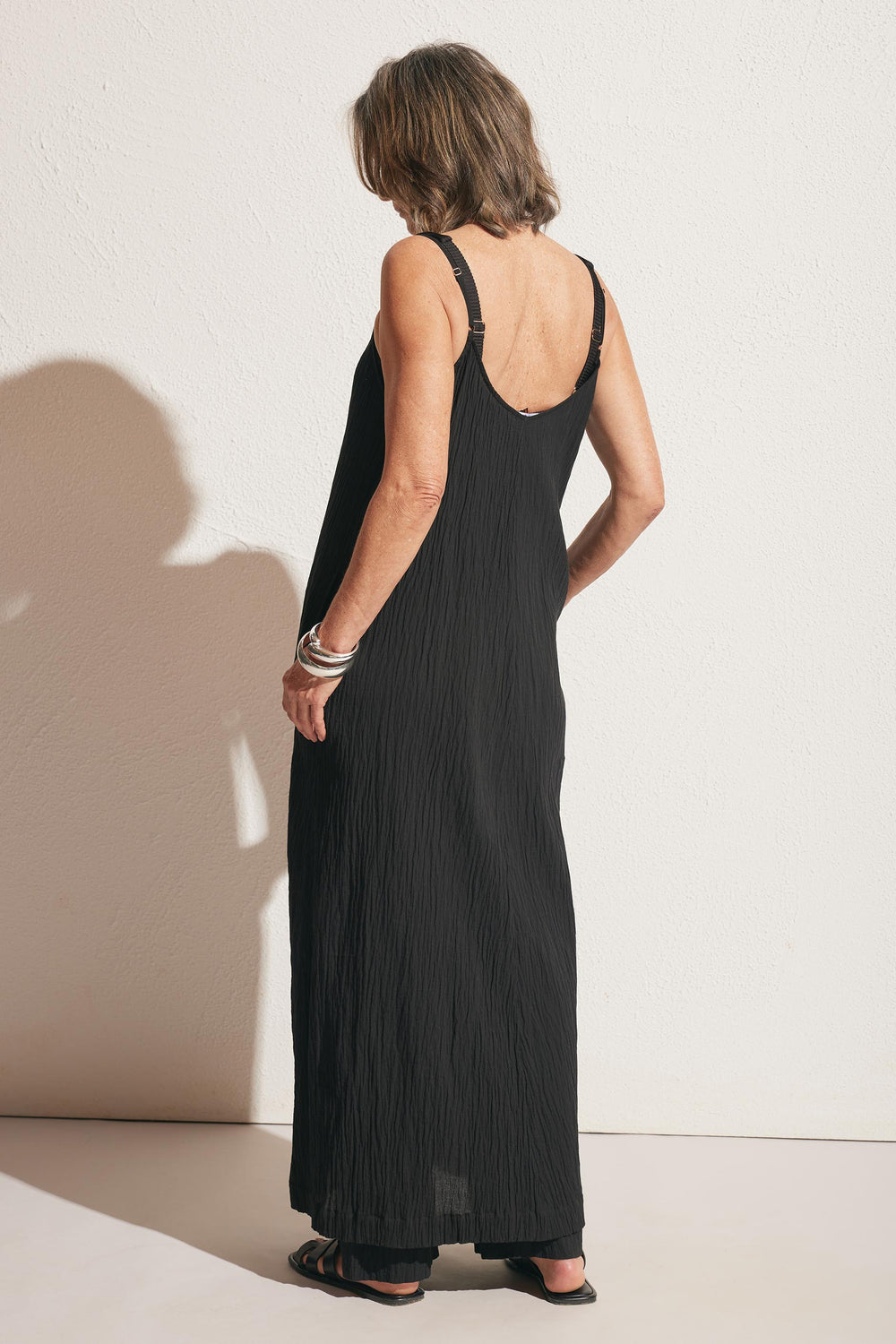 Woman wearing a sleeveless black maxi dress with textured fabric, accessorized with silver bracelets and black sandals, shown from the back