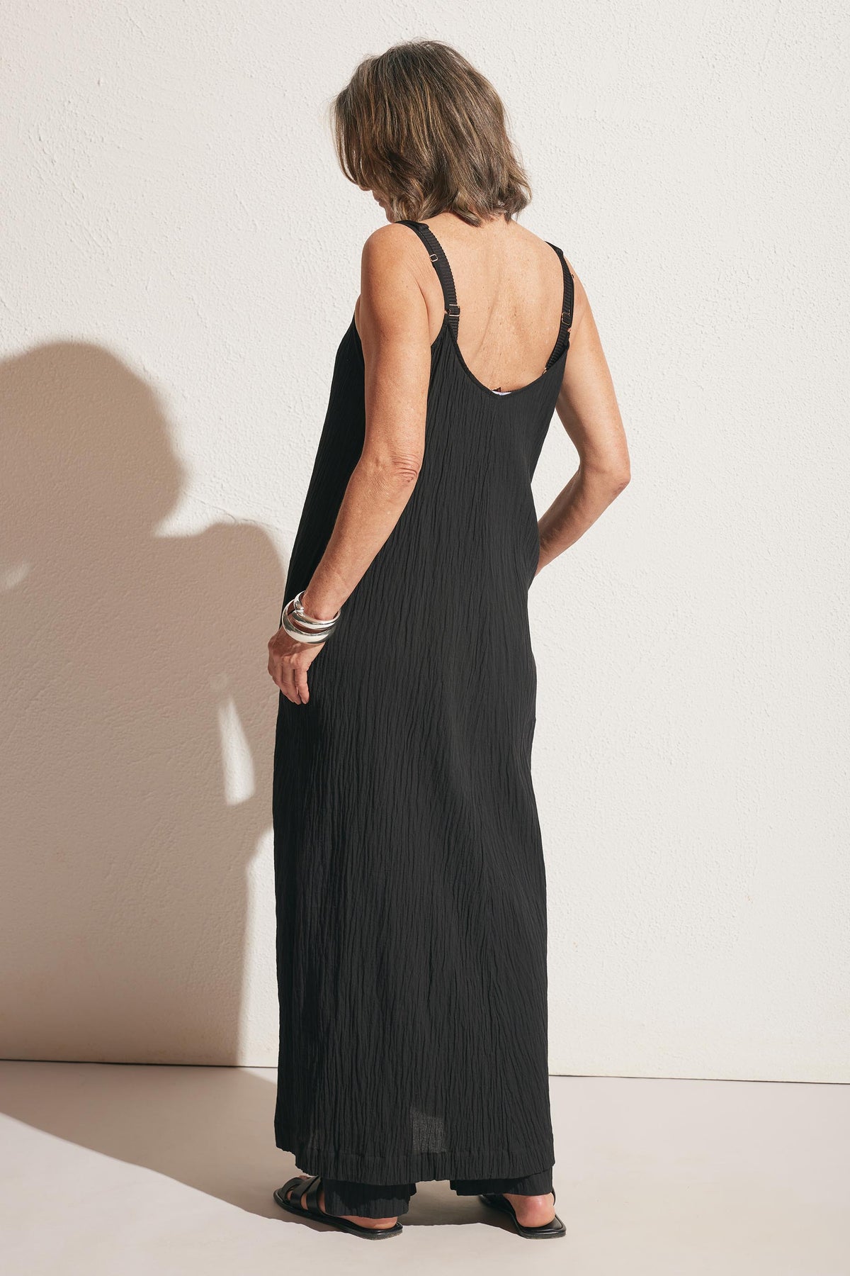 Woman wearing a sleeveless black maxi dress with textured fabric, accessorized with silver bracelets and black sandals, shown from the back