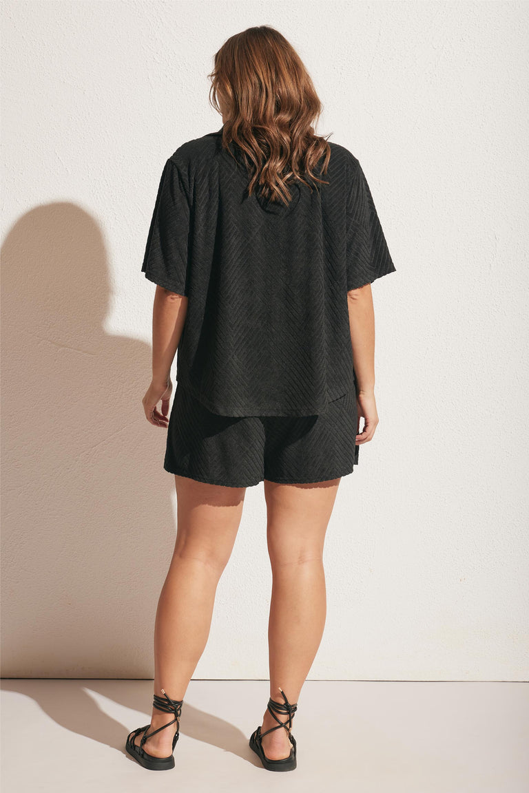 Woman wearing relaxed black textured shorts and matching oversized short-sleeve top with lace-up sandals, facing away