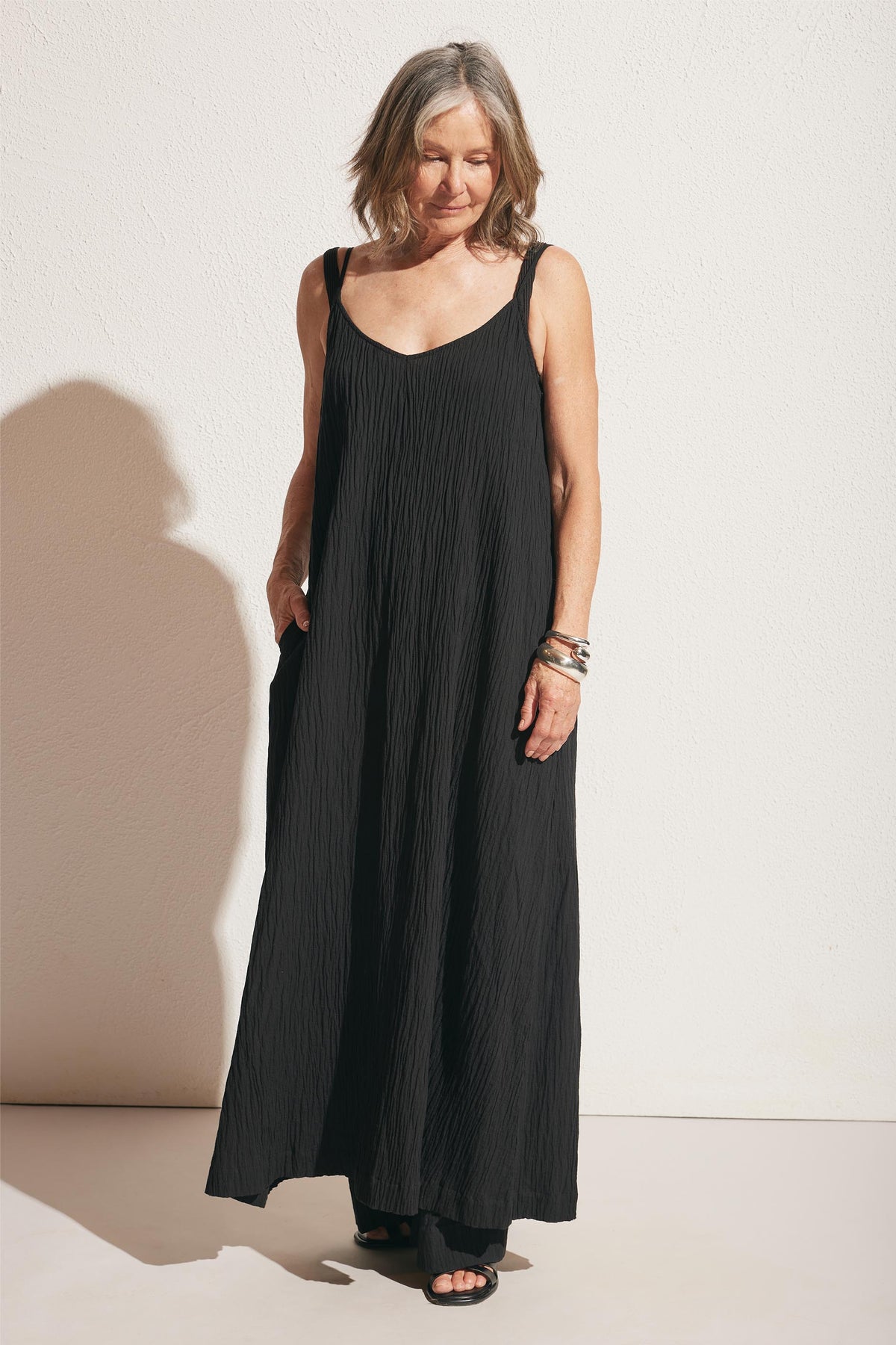 Woman wearing a relaxed black maxi dress with thin straps and subtle textured fabric, styled with silver bangles and black sandals
