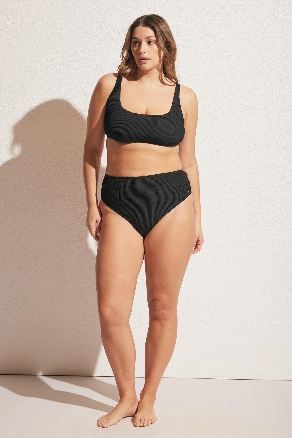 Woman wearing a textured black high-waisted bikini bottom and matching scoop neck top standing against a light wall