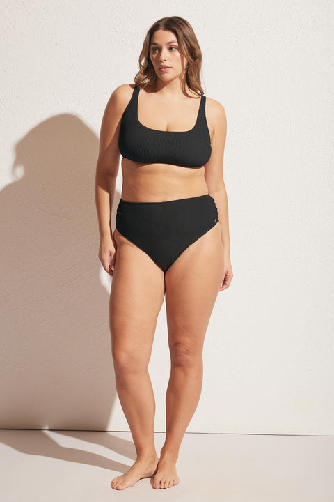 Woman wearing a textured black high-waisted bikini bottom and matching scoop neck top standing against a light wall
