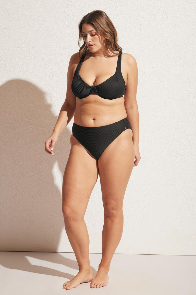 Woman wearing a black ribbed underwire bikini top and mid-rise bikini bottoms, standing barefoot against a light wall.