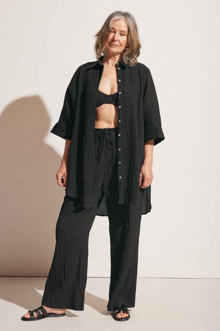 Older woman wearing relaxed black crinkled pants and a matching long button-up shirt with black sandals