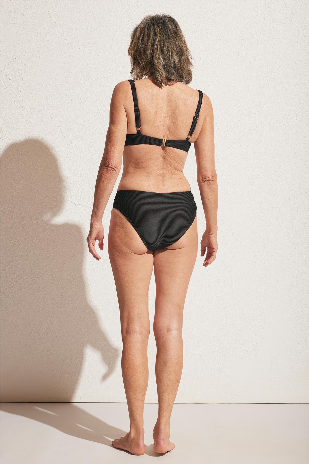 Older woman shown from behind wearing a black ribbed bikini top with adjustable straps and matching bikini bottoms