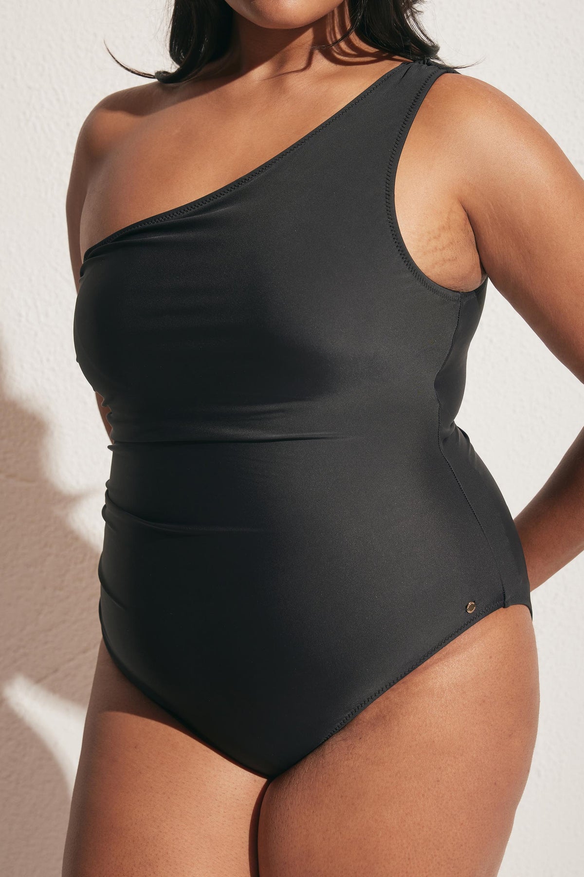 Close-up of woman wearing a matte black one-shoulder swimsuit with clean lines against a light textured wall