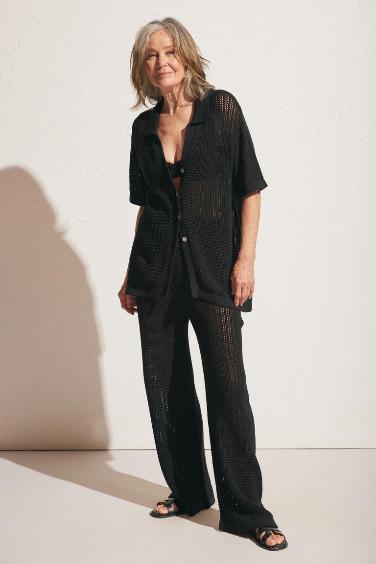 Mature woman wearing a black open-knit button-up top and matching pants with sandals, standing in soft sunlight.