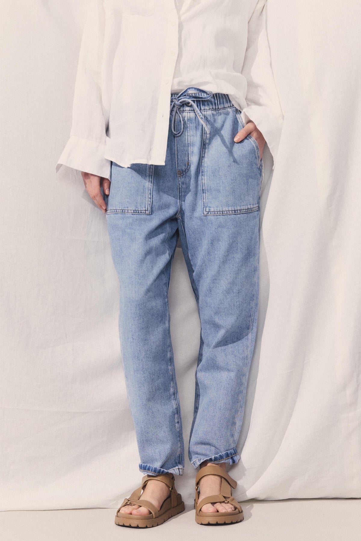 Model wearing high rise cropped blue denim jogger pants with an elastic drawstring waist and functional pockets