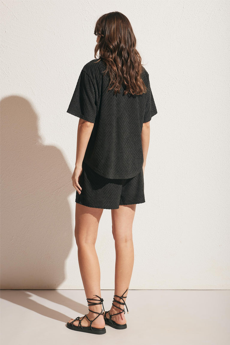 Woman wearing a relaxed black textured short and matching loose short-sleeve top with lace-up sandals, seen from behind