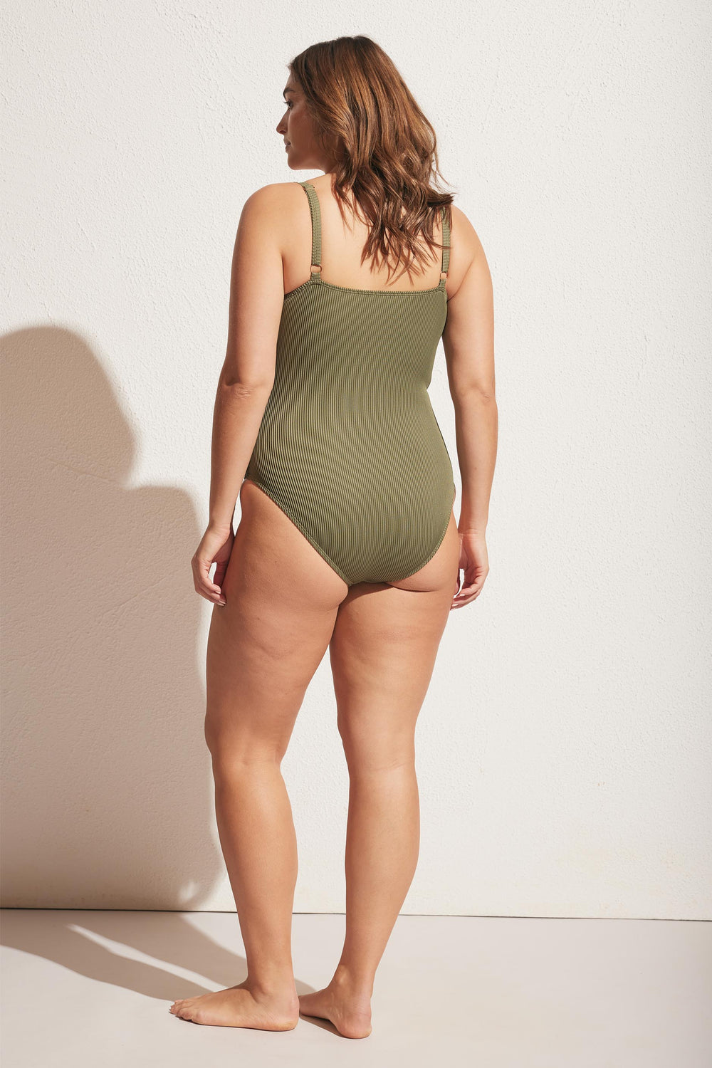 Woman wearing a soft olive ribbed swimsuit with adjustable straps, shown from the back in a relaxed pose