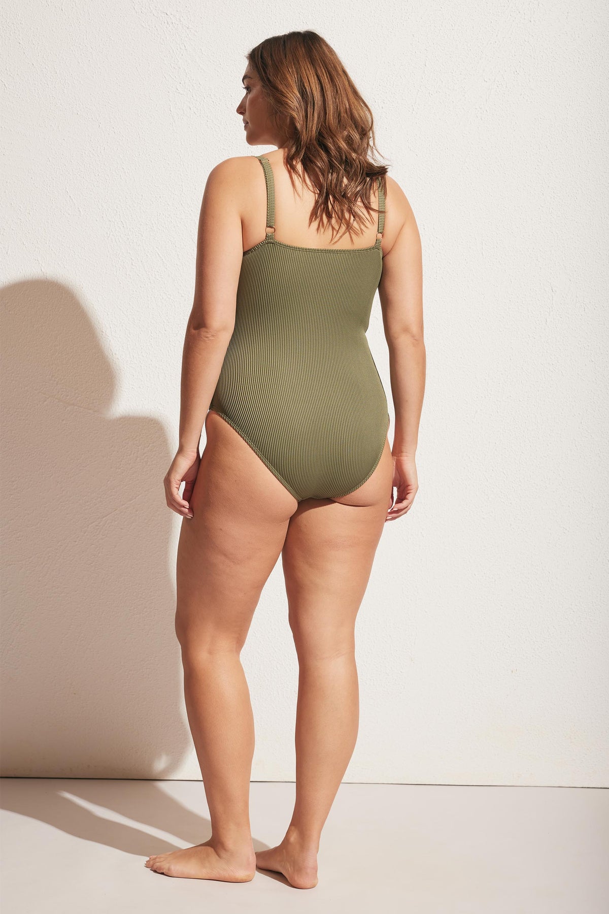 Woman wearing a soft olive ribbed swimsuit with adjustable straps, shown from the back in a relaxed pose