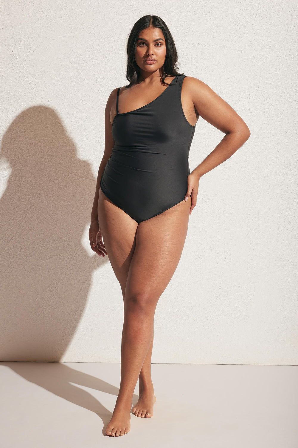 Woman wearing a sleek black matte one shoulder swimsuit standing barefoot against a textured light wall background