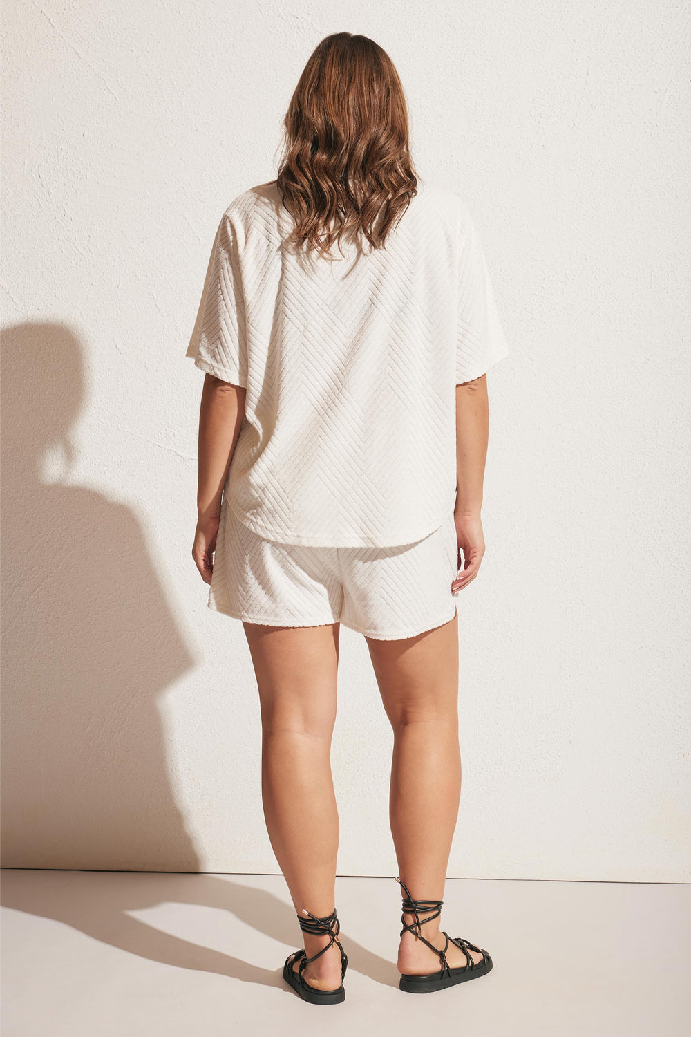 Woman wearing a relaxed white textured short set with loose sleeves, paired with black lace-up sandals, standing against a wall