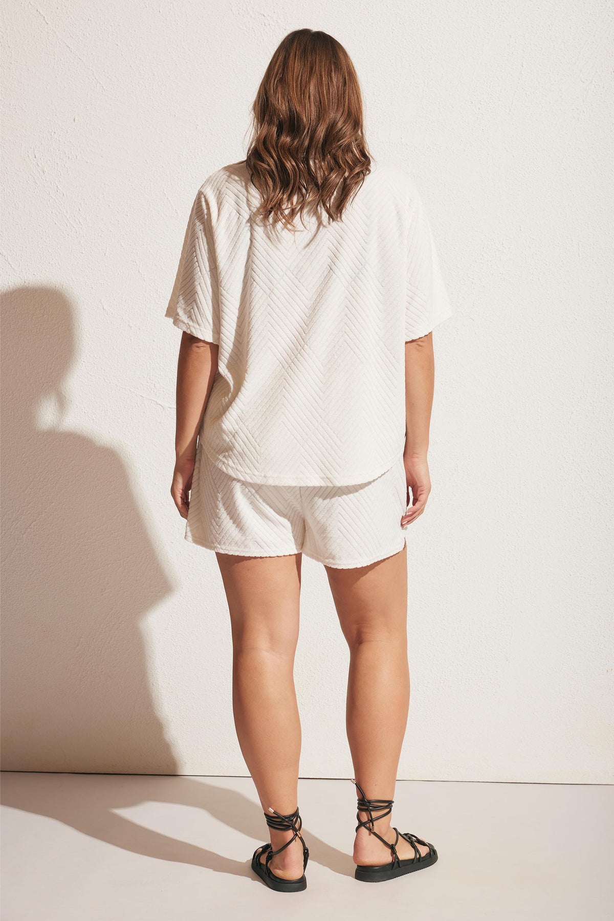 Woman wearing a relaxed white textured short set with loose sleeves, paired with black lace-up sandals, standing against a wall