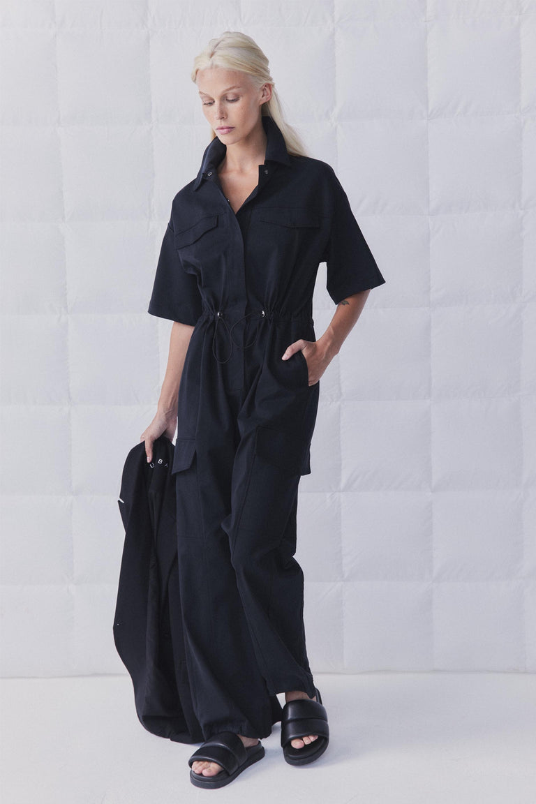 Woman wearing a black collared cargo jumpsuit with short sleeves, elasticated waist, snap front and utility pockets