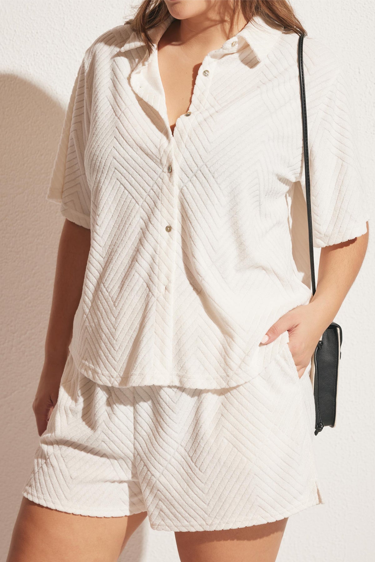 Woman wearing textured cream relaxed-fit shorts and matching short-sleeve button-up top with a black crossbody bag