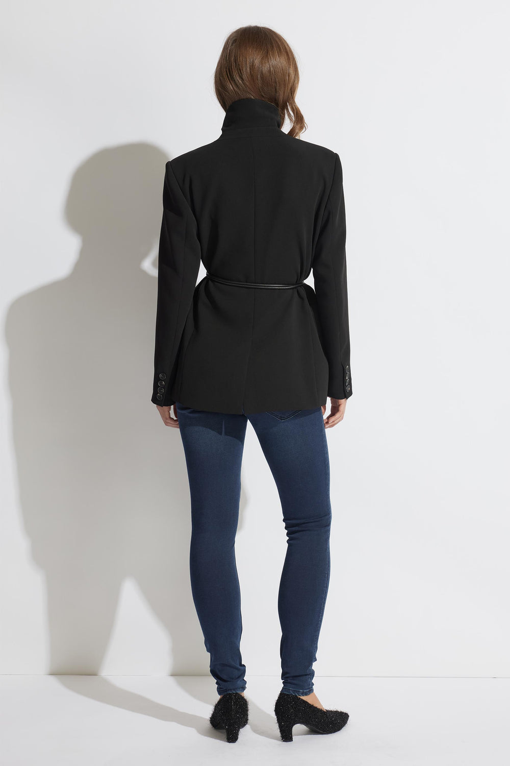 Woman wearing a black belted blazer paired with dark blue skinny jeans and black heeled shoes standing against a white wall