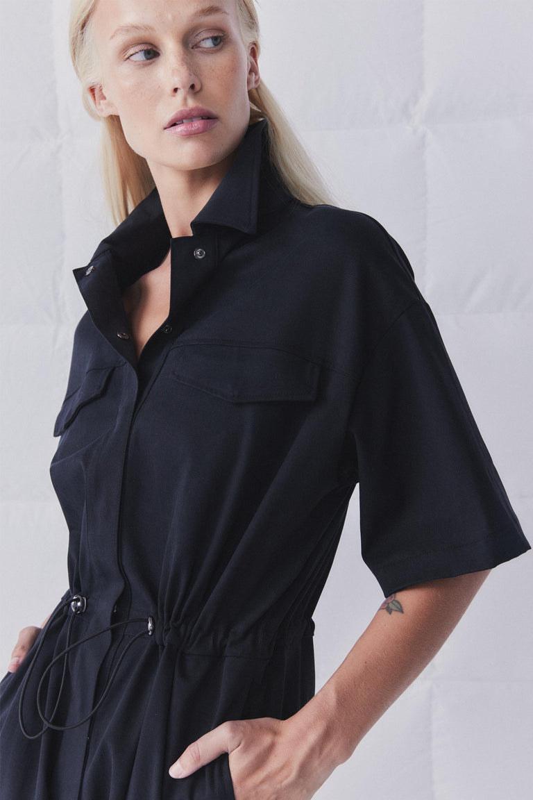 Woman wearing a black collared jumpsuit with short sleeves, snap front, chest pockets and adjustable drawstring waist