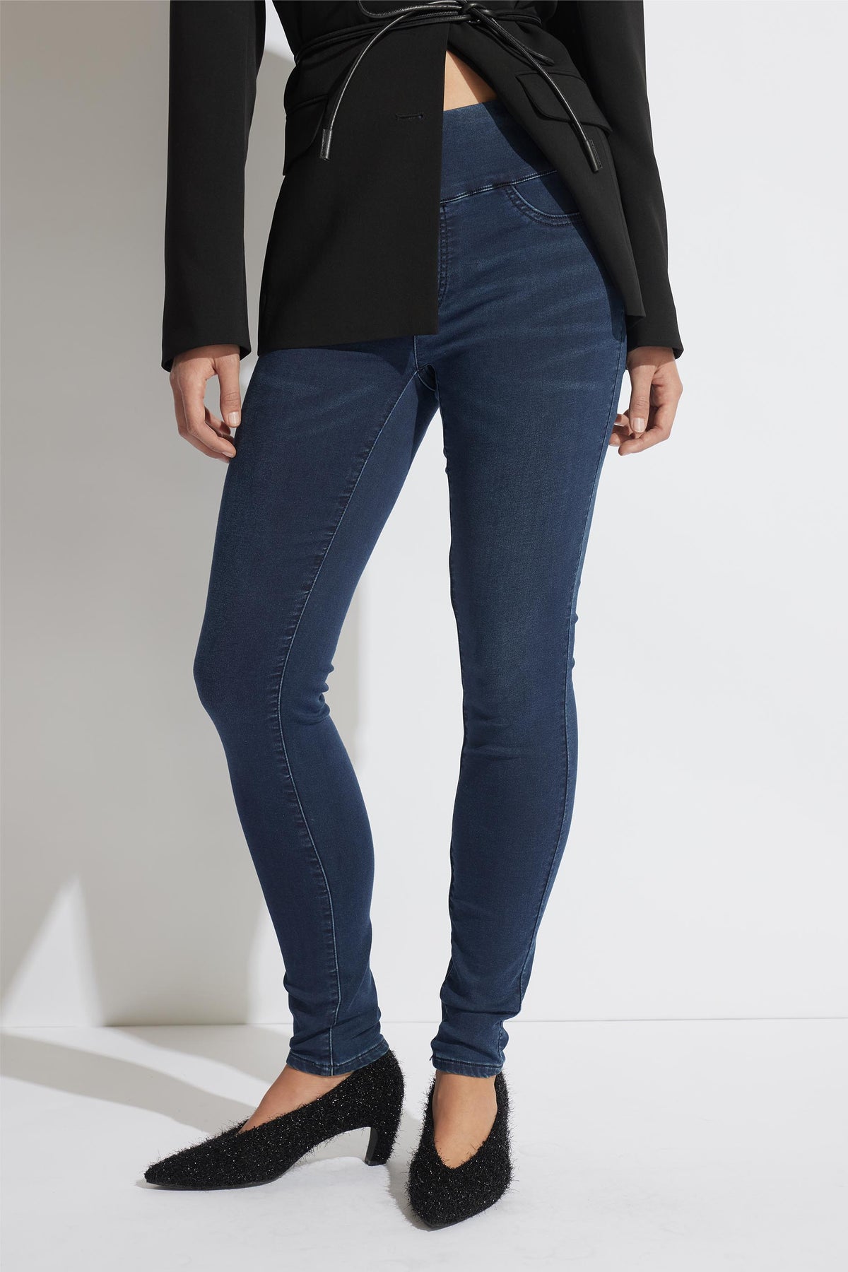 Woman wearing dark indigo skinny jeans paired with a tailored black blazer and textured black heels against a minimalist background