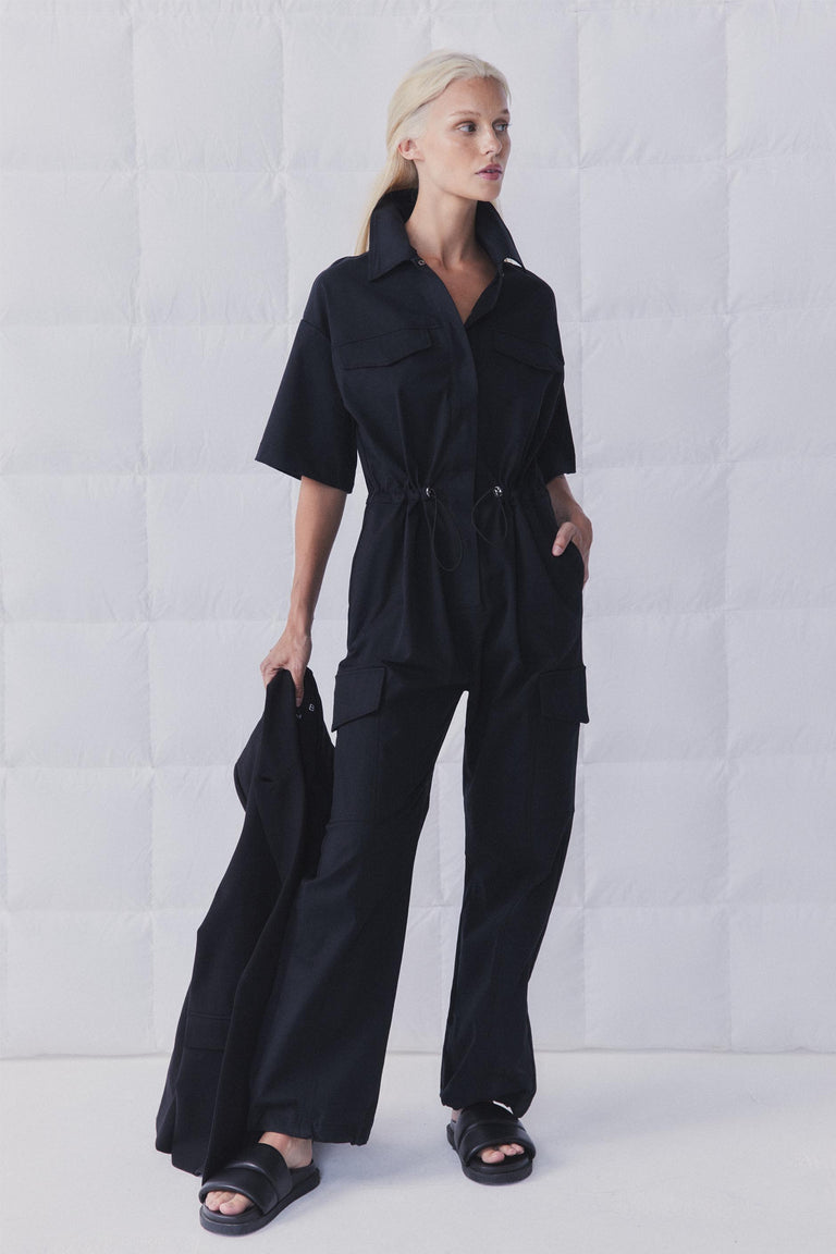 Woman wearing a black collared cargo jumpsuit with short sleeves, an elasticated waist and wide full length legs