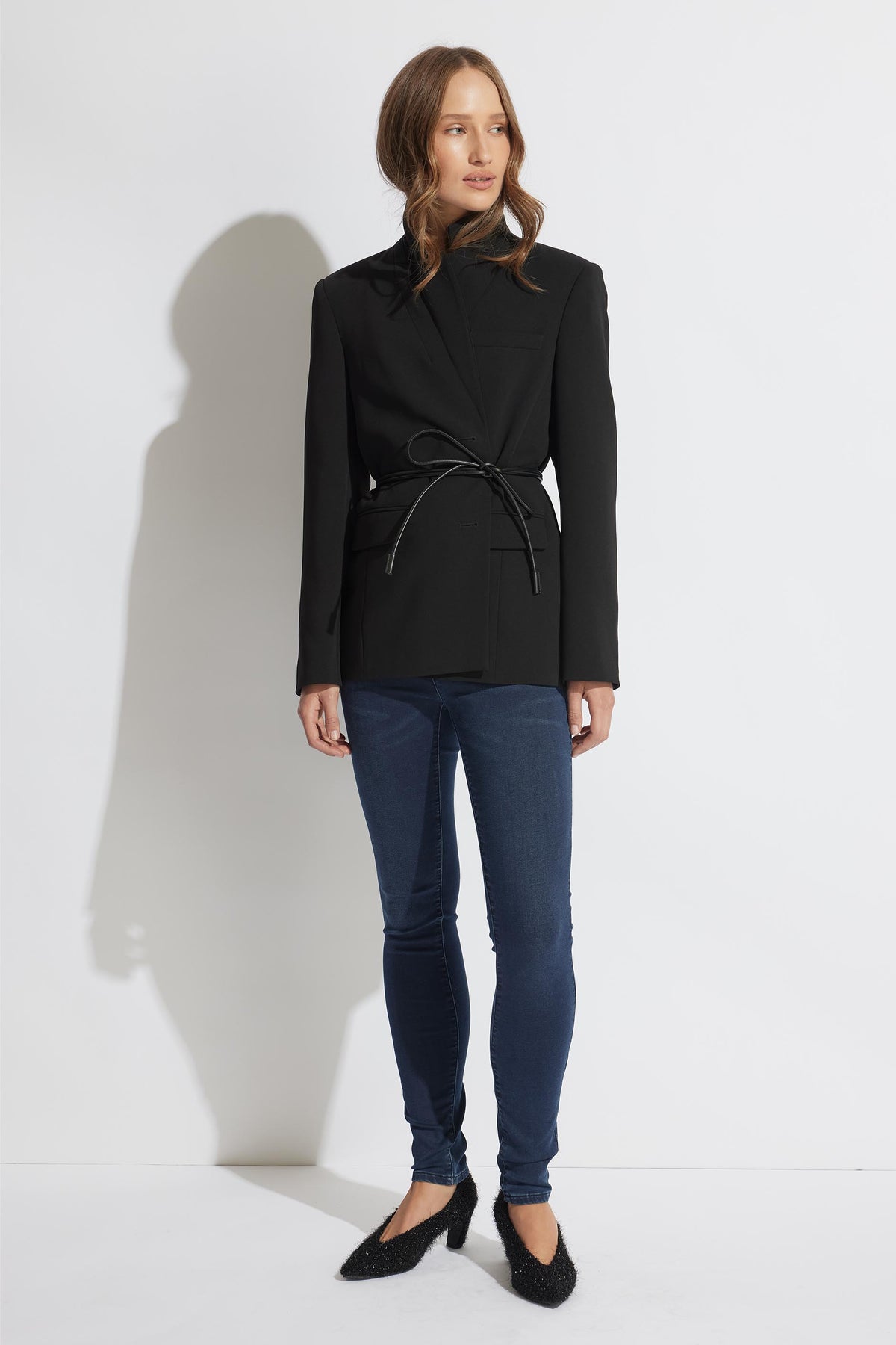 Woman wearing a black belted blazer paired with dark blue skinny jeans and textured black pointed flats against a white background