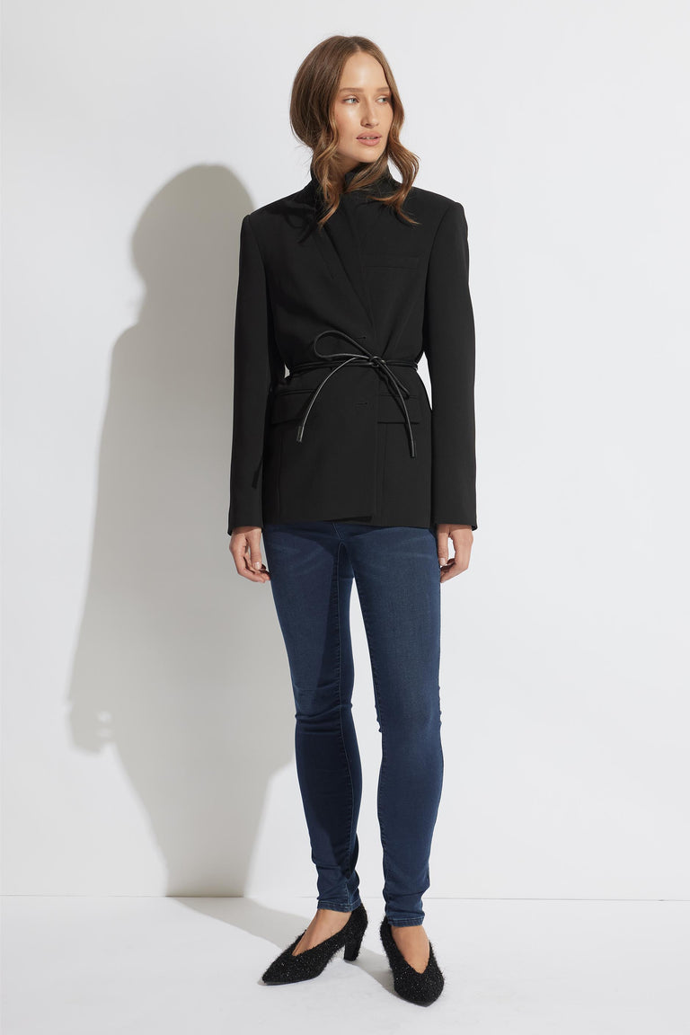 Woman wearing a black belted blazer paired with dark blue skinny jeans and textured black pointed flats against a white background