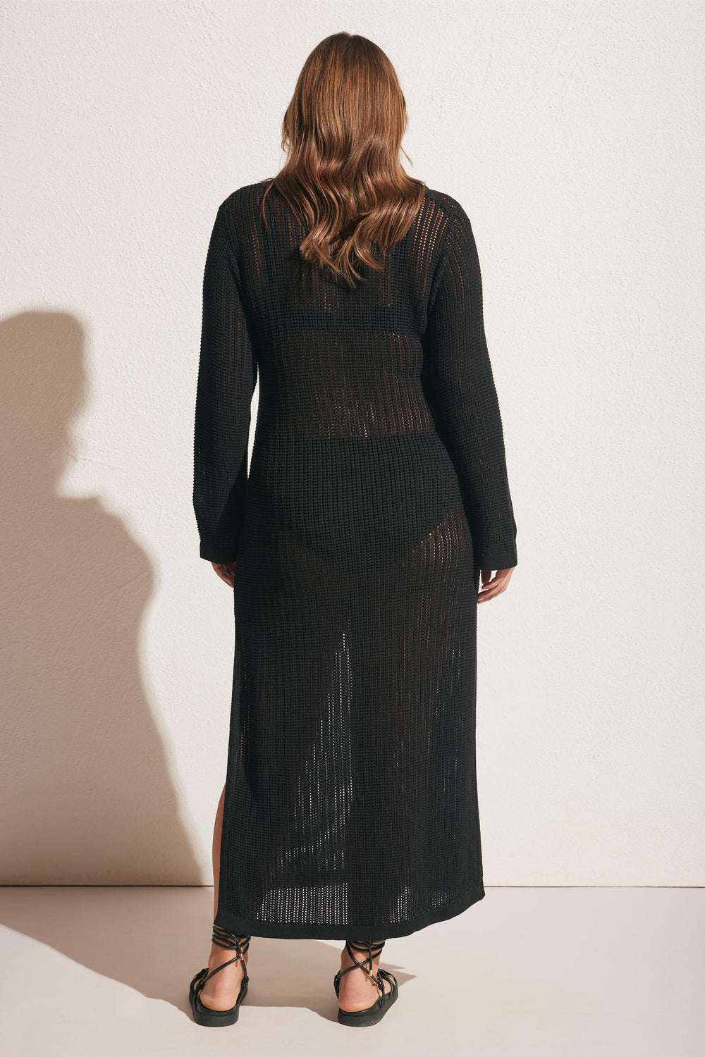 Woman seen from behind wearing a long black crochet dress with long sleeves and flat lace-up sandals against a plain wall