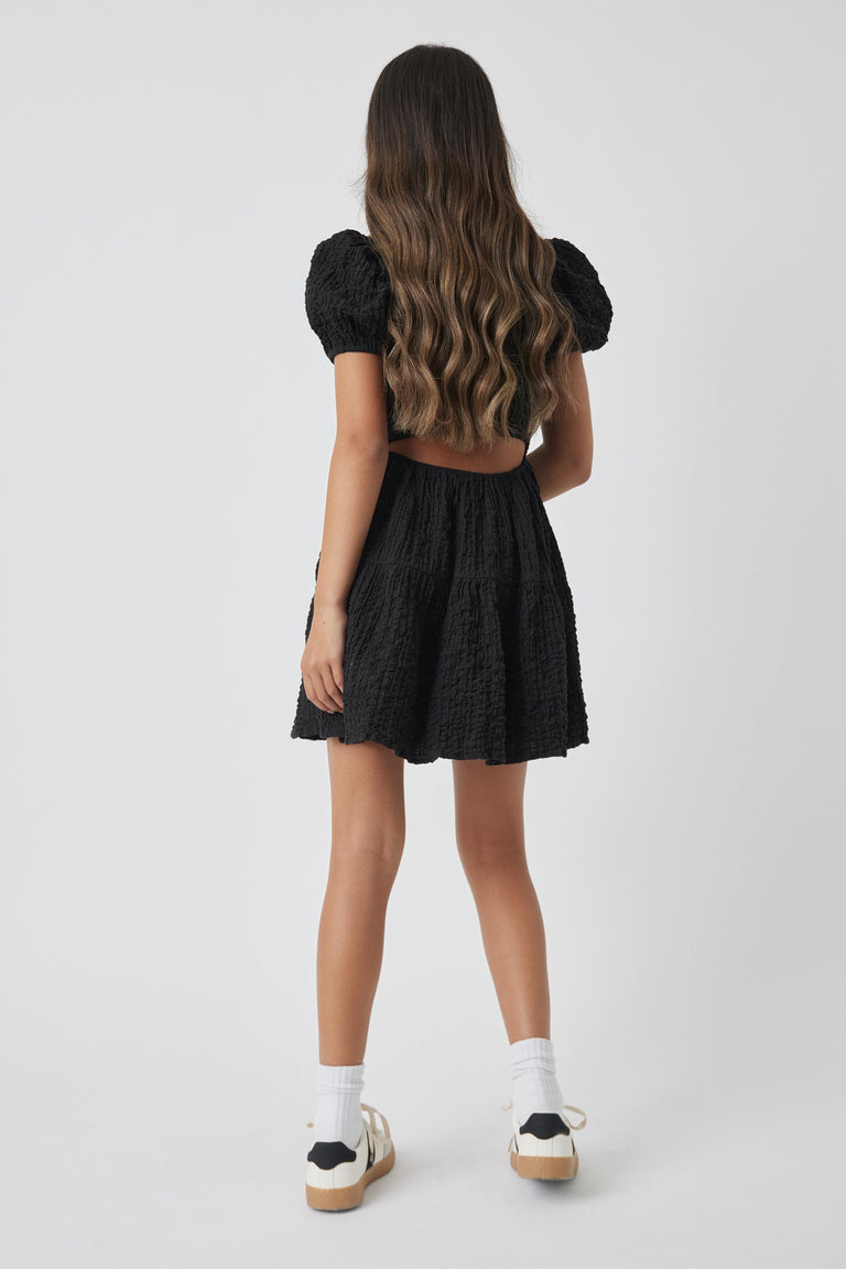 Girl wearing a black textured mini dress with puff sleeves and open back, paired with white socks and sneakers.