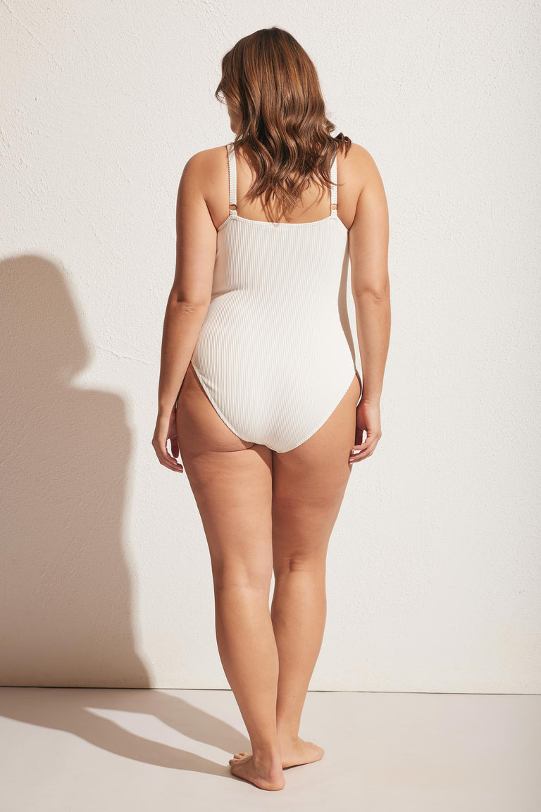 Woman wearing an off-white ribbed one-piece with adjustable straps, shown from the back against a neutral wall.