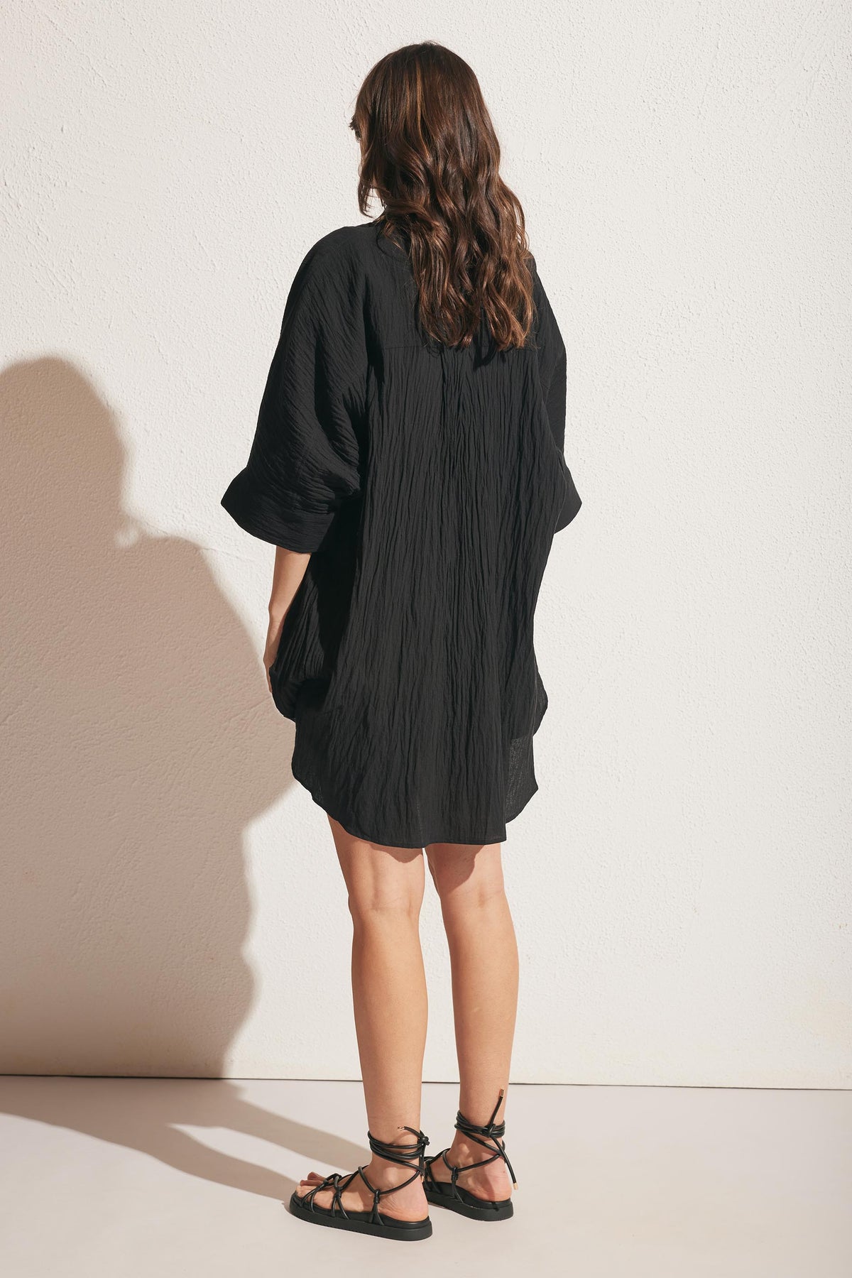 Woman with long wavy hair wearing a loose black textured dress and black lace-up sandals against a light wall