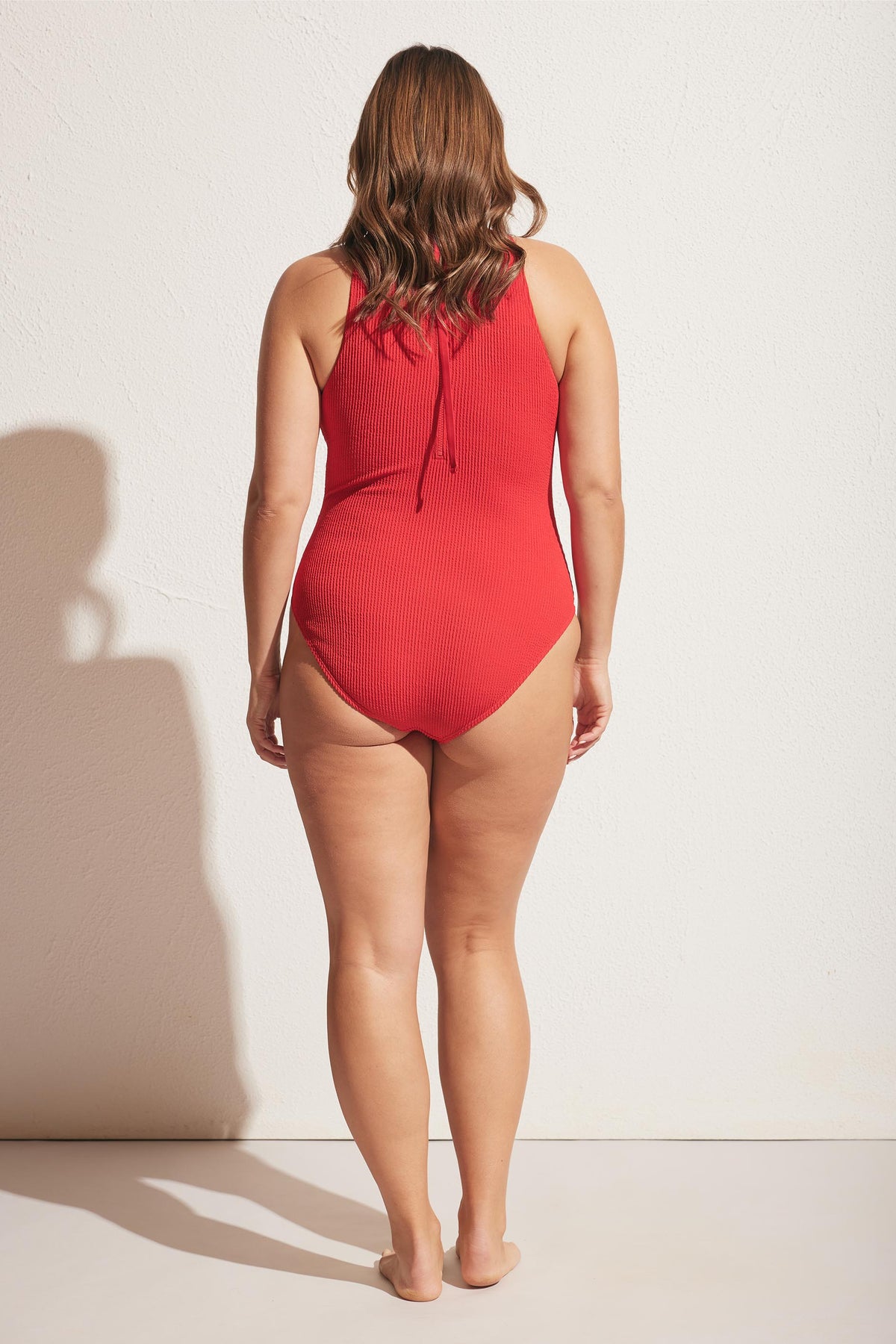 Woman shown from behind wearing a red textured racerback one-piece swimsuit against a neutral wall