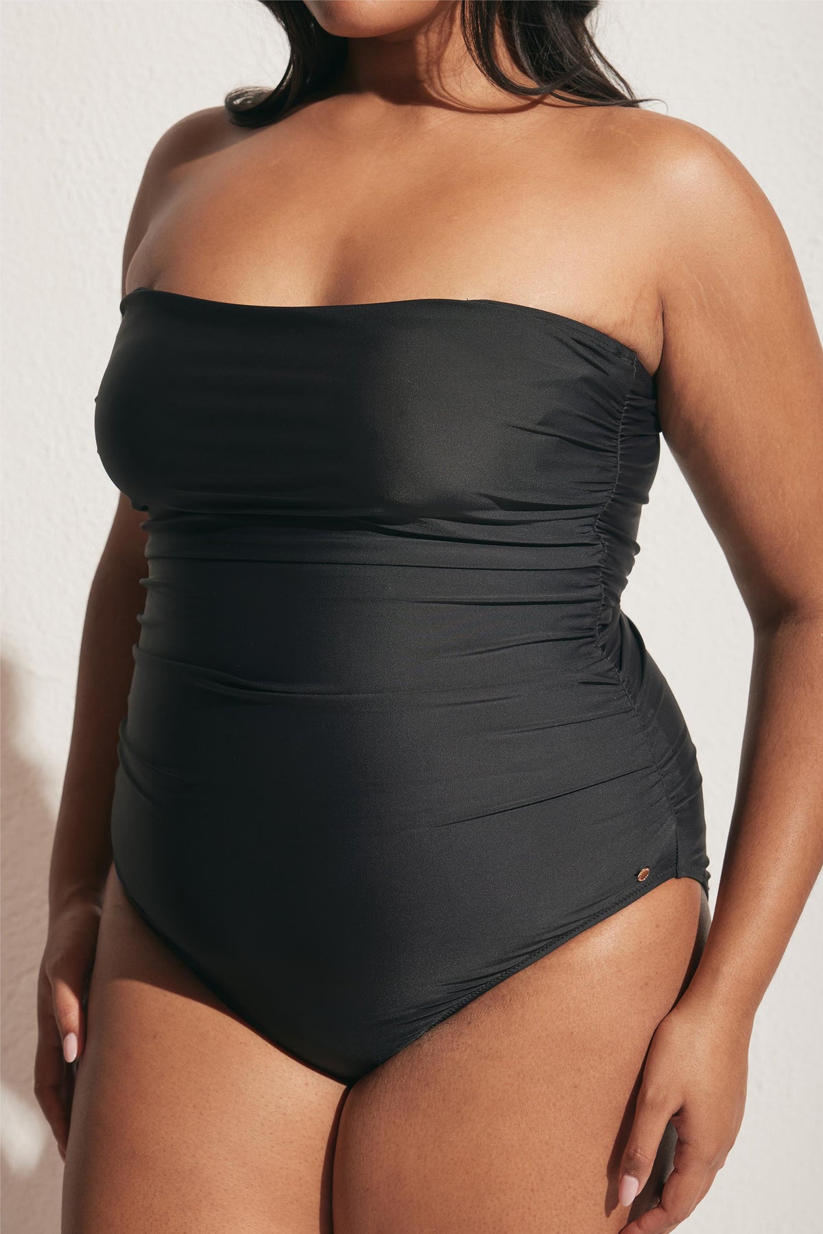 Close-up of a woman wearing a black matte ruched strapless one-piece swimsuit in natural sunlight against a white background
