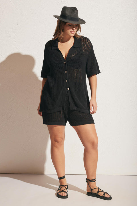 Woman wearing a black crochet button-up top and matching shorts with strappy black sandals and a grey fedora hat