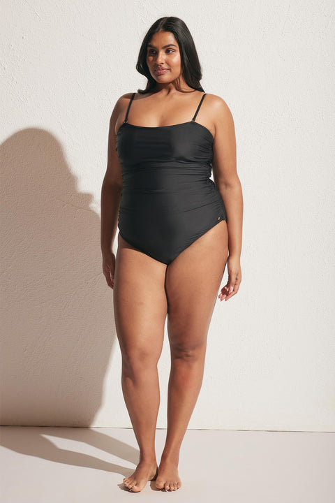 Model wearing a black matte one-piece swimsuit with thin straps and subtle ruching, standing barefoot against a textured wall.