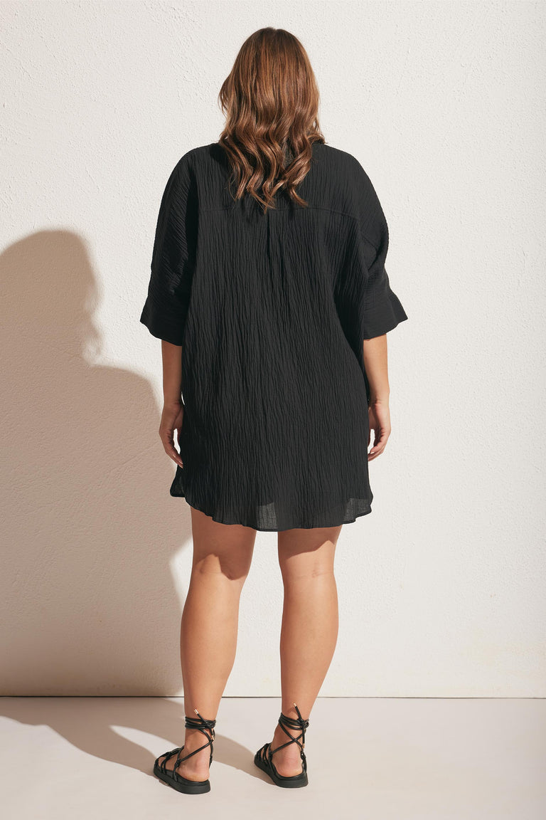 Woman wearing a loose black textured beach cover-up dress with rolled sleeves and black lace-up sandals, back view