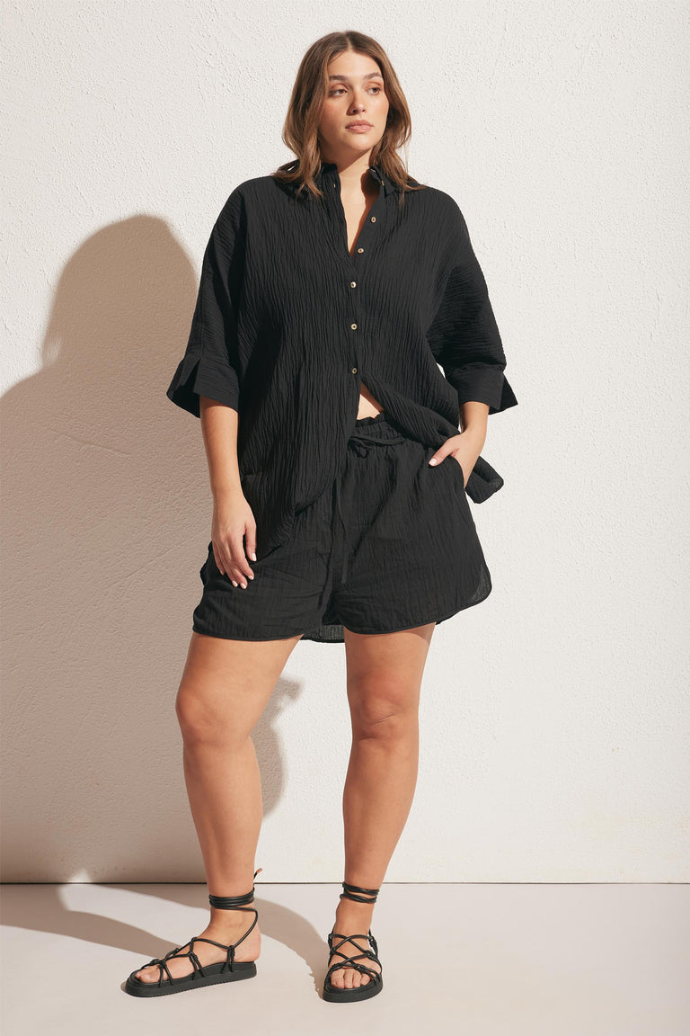 Woman wearing relaxed black textured button-up shirt and matching shorts with lace-up flat sandals against light wall