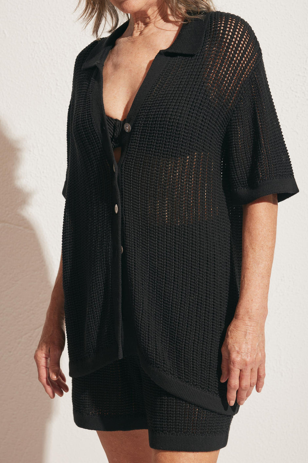 Woman wearing a black open-knit short-sleeve shirt over matching shorts and a black bandeau top against a light wall