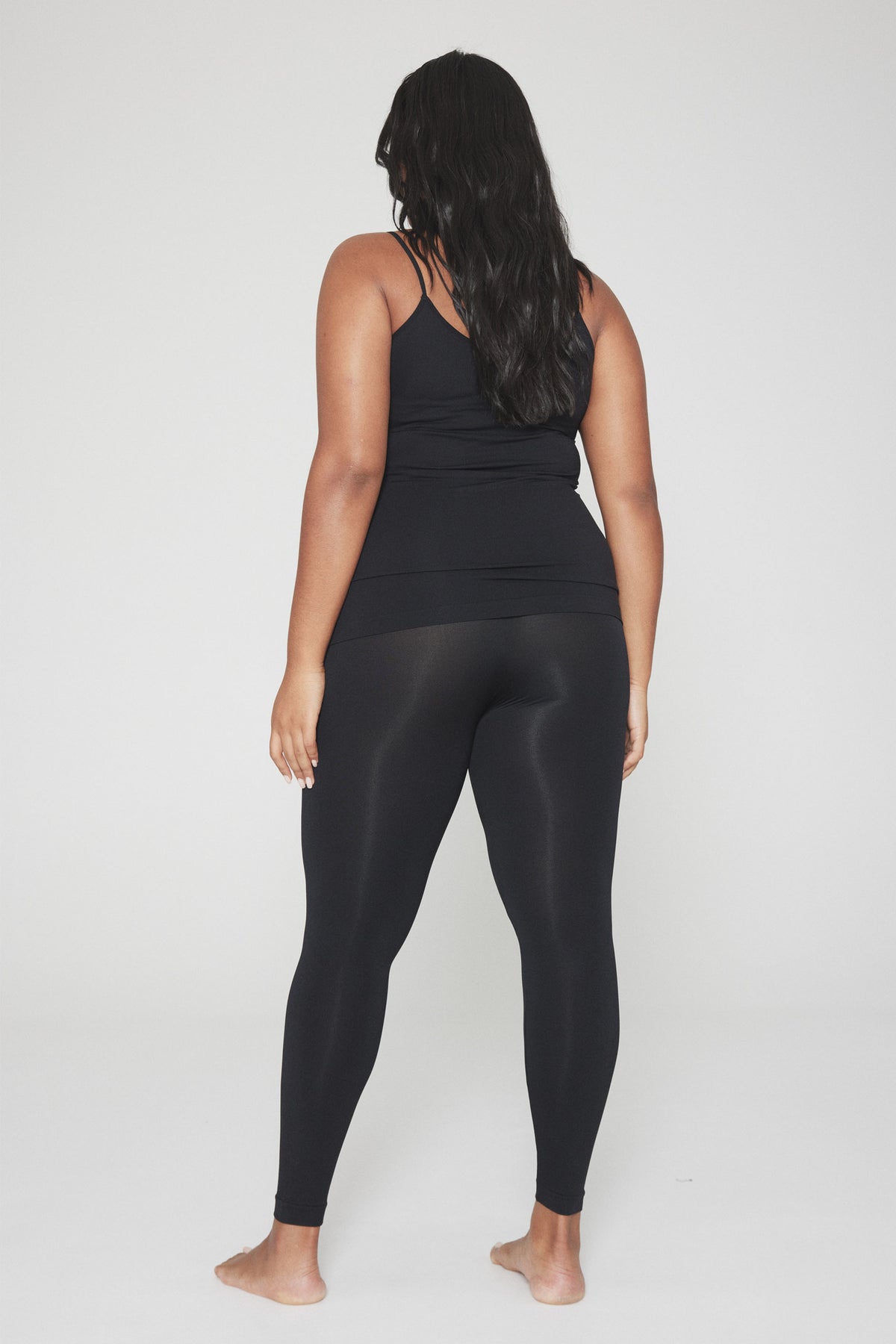 Woman seen from behind wearing fitted black seamless leggings and a matching black tank top, standing barefoot.