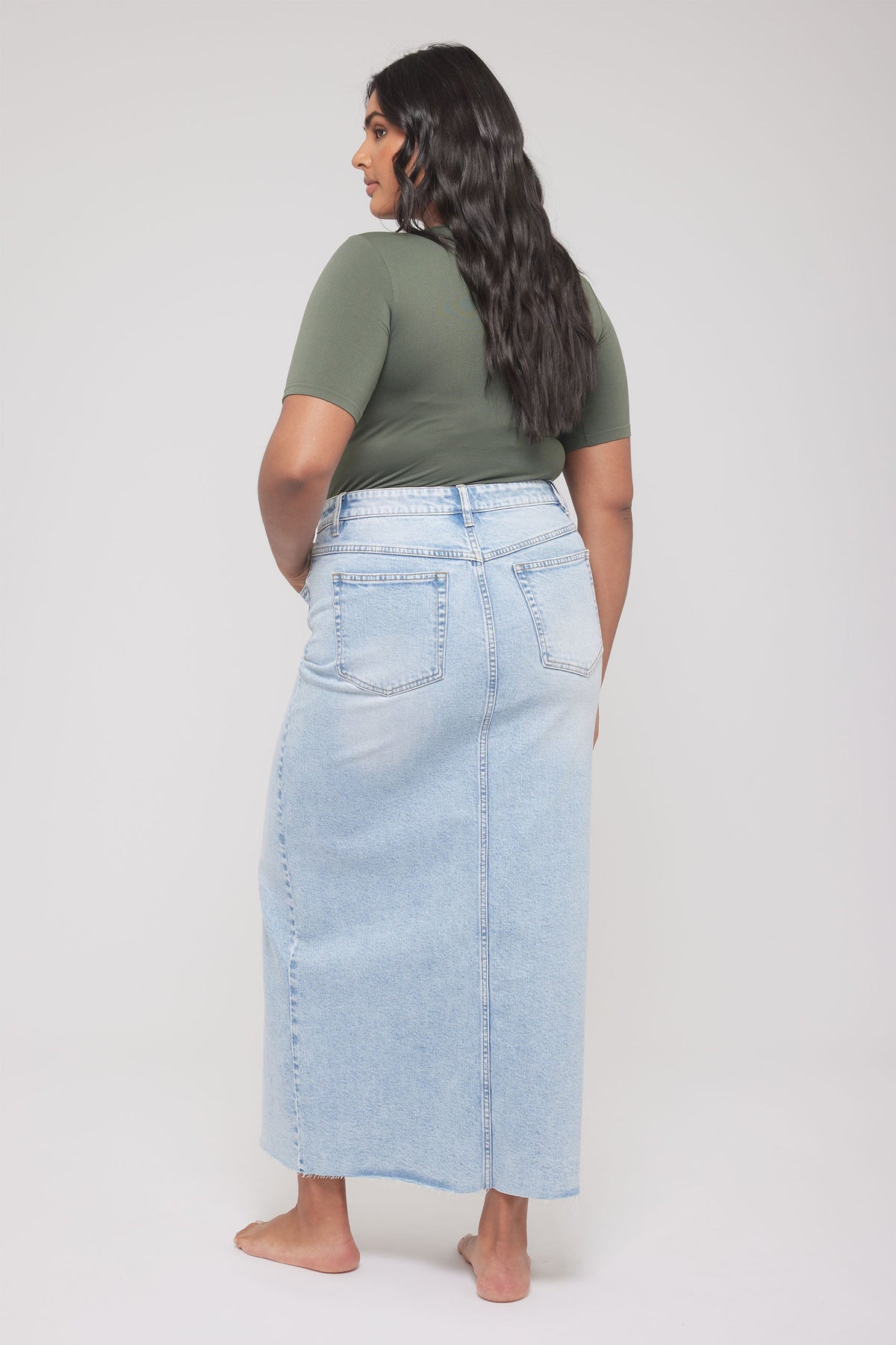 Woman with long dark hair wearing an olive fitted tee paired with a light blue high-waisted denim skirt barefoot