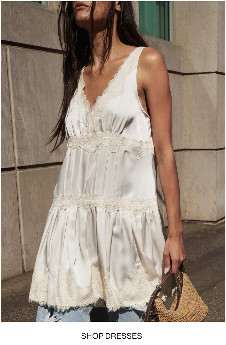 A woman wearing a white satin sleeveless dress with lace trim, standing outdoors against a stone wall and holding a woven straw bag. The text 'SHOP DRESSES' appears at the bottom.