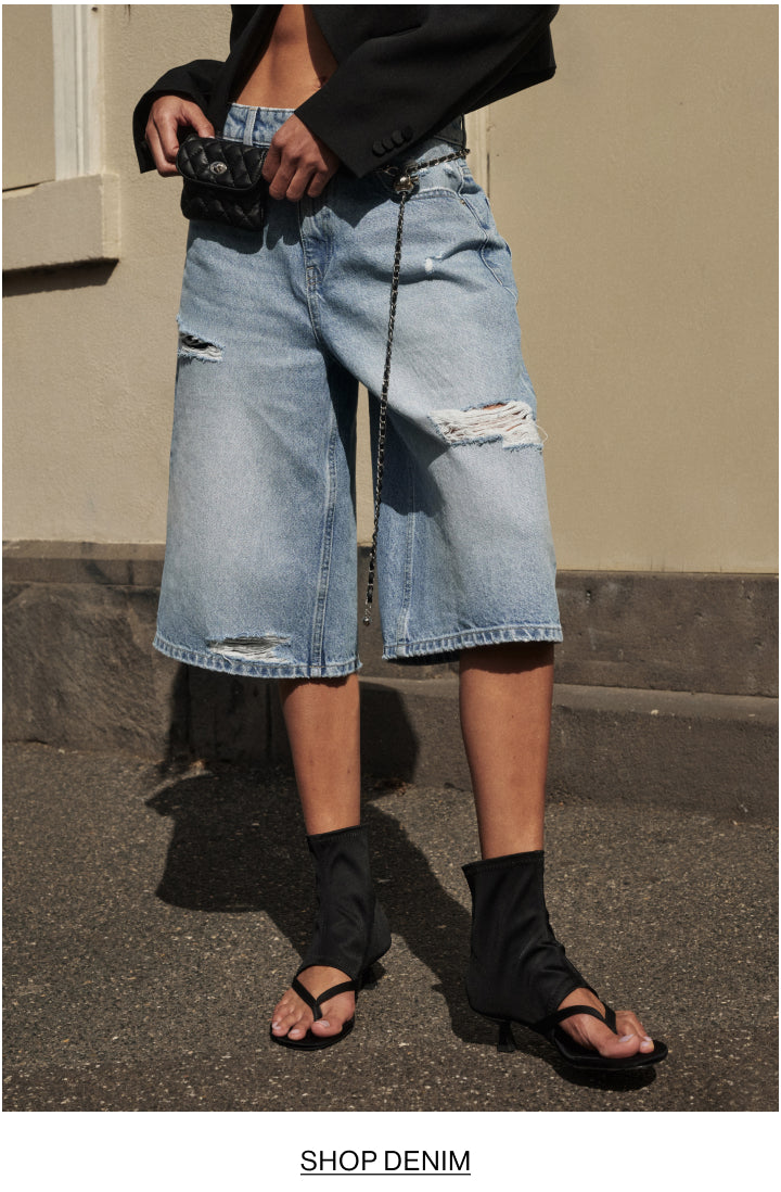 A person wearing light-wash distressed denim shorts with ripped details, styled with a black blazer, black ankle socks, and black sandals, standing outdoors against a beige wall.