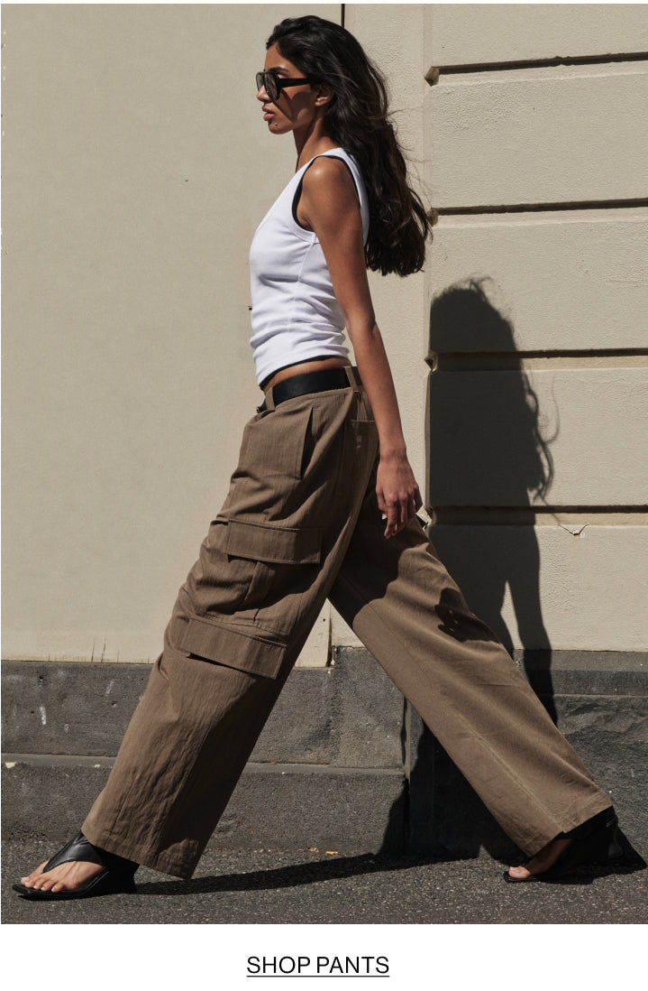 A woman walking outdoors in profile wearing oversized brown cargo pants, a white tank top, black sandals, and sunglasses against a beige wall.